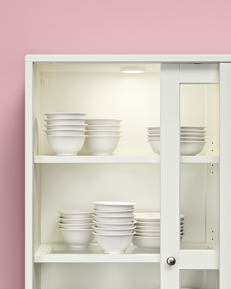 A white HAVSTA sliding-glass-door storage combination with stacked white bowls and plates inside, set against a pink wall.