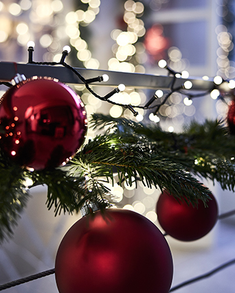Una cadena luminosa STRÅLA para exteriores, una guirnalda artificial verde VINTERFINT y grandes adornos rojos para el árbol colgados en la barandilla del balcón.