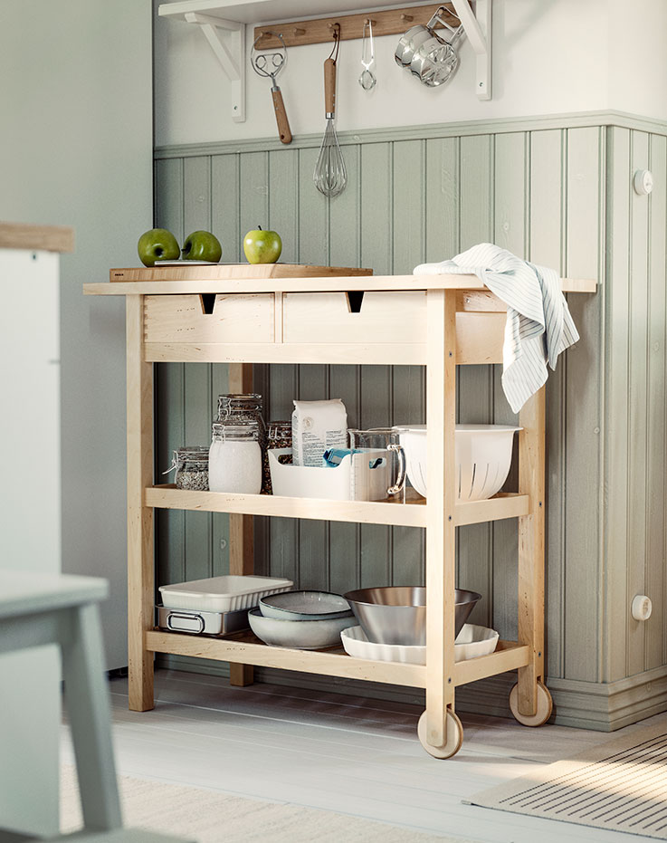 Un carrito de cocina de abedul de la marca FÖRHÖJA, completamente cargado, aparcado junto a una pared revestida de paneles de color verde salvia en una cocina.
