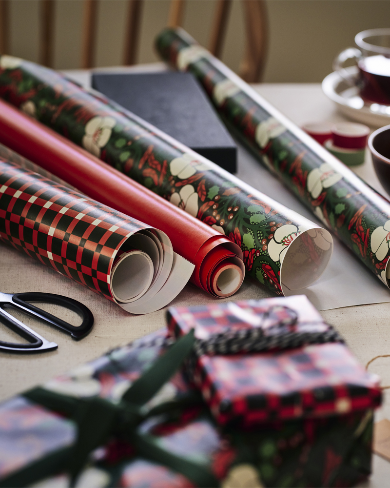 Rollos de papel de regalo navideño con motivos florales y a cuadros, tijeras y regalos envueltos sobre una mesa.