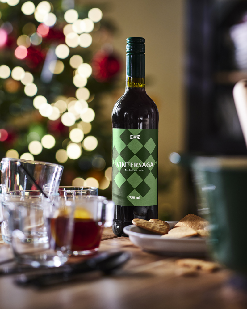 Botella de bebida de frutas VINTERSAGA de IKEA junto a vasos y galletas con luces navideñas desenfocadas de fondo.