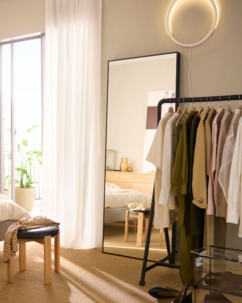 Bright bedroom corner with HOVET mirror, TURBO clothes rack, VARMBLIXT wall lamp and DYTÅG sheer curtains.