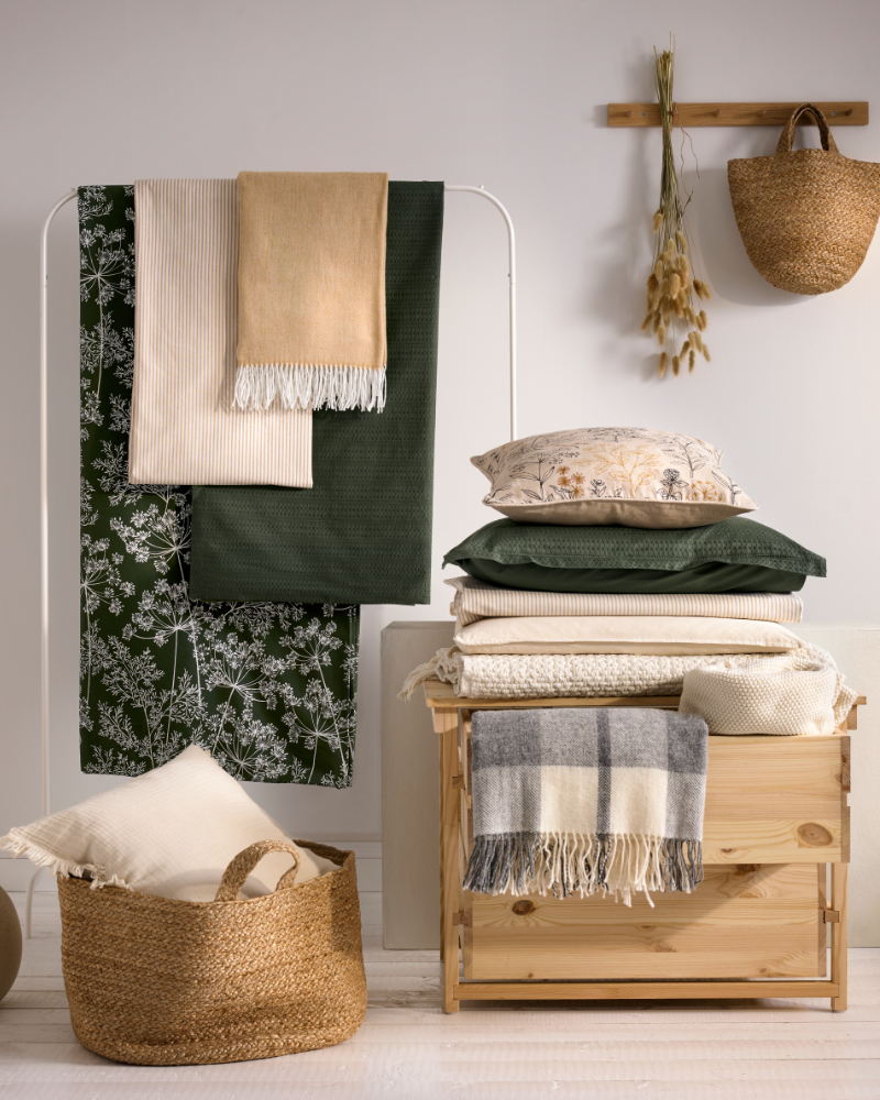 Riel blanco con mantas colgadas, mueble de madera con textiles doblados y cesta decorativa.
