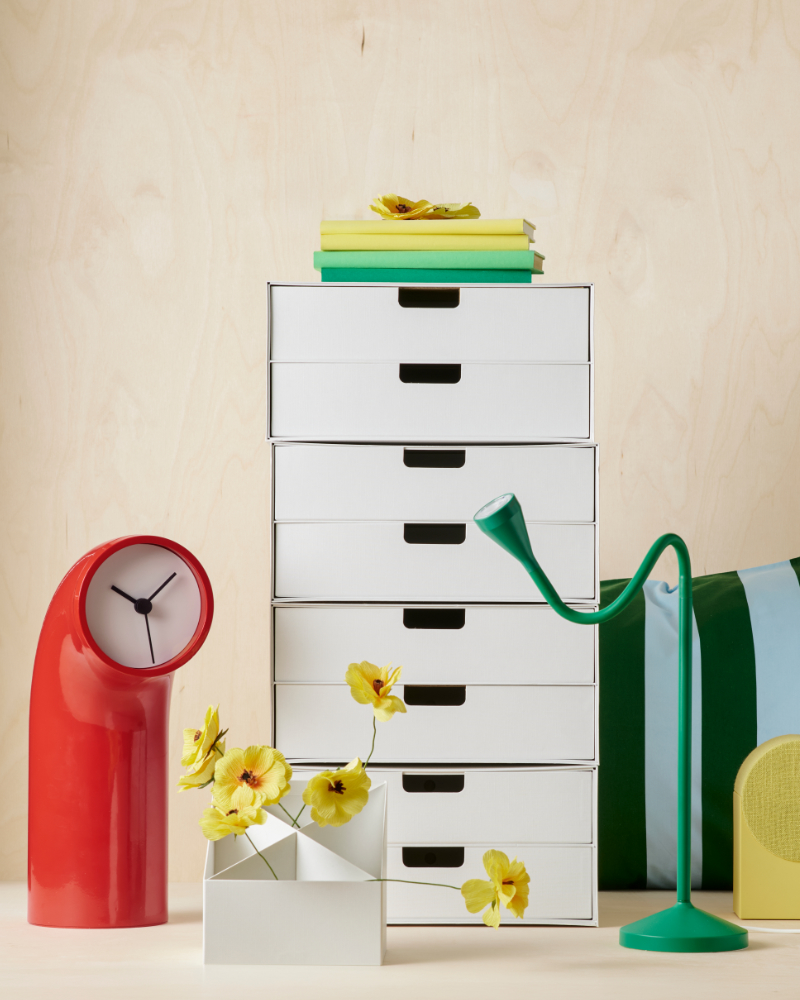 Organizador blanco con flores amarillas, reloj rojo, lámpara verde y libros.