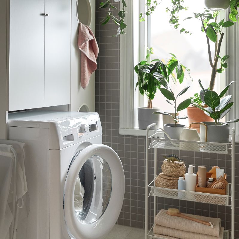 Un baño funcional con azulejos grises, lavadora, lavabo y plantas junto a una ventana. Ambiente ordenado y luminoso.