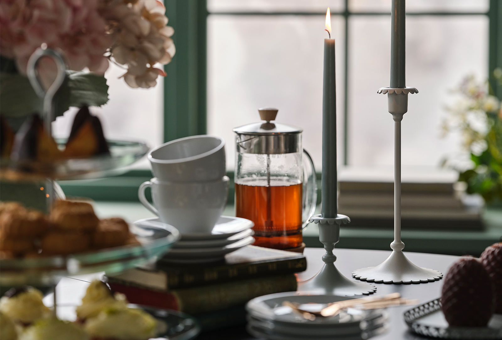 Dos portavelas SNÖSISKA de color blanco roto están sobre una mesa de comedor, con té y pasteles delante de una ventana verde.