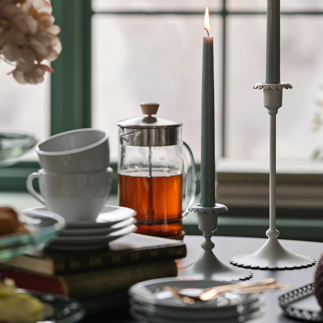 Dos portavelas SNÖSISKA de color blanco roto están sobre una mesa de comedor, con té y pasteles delante de una ventana verde.
