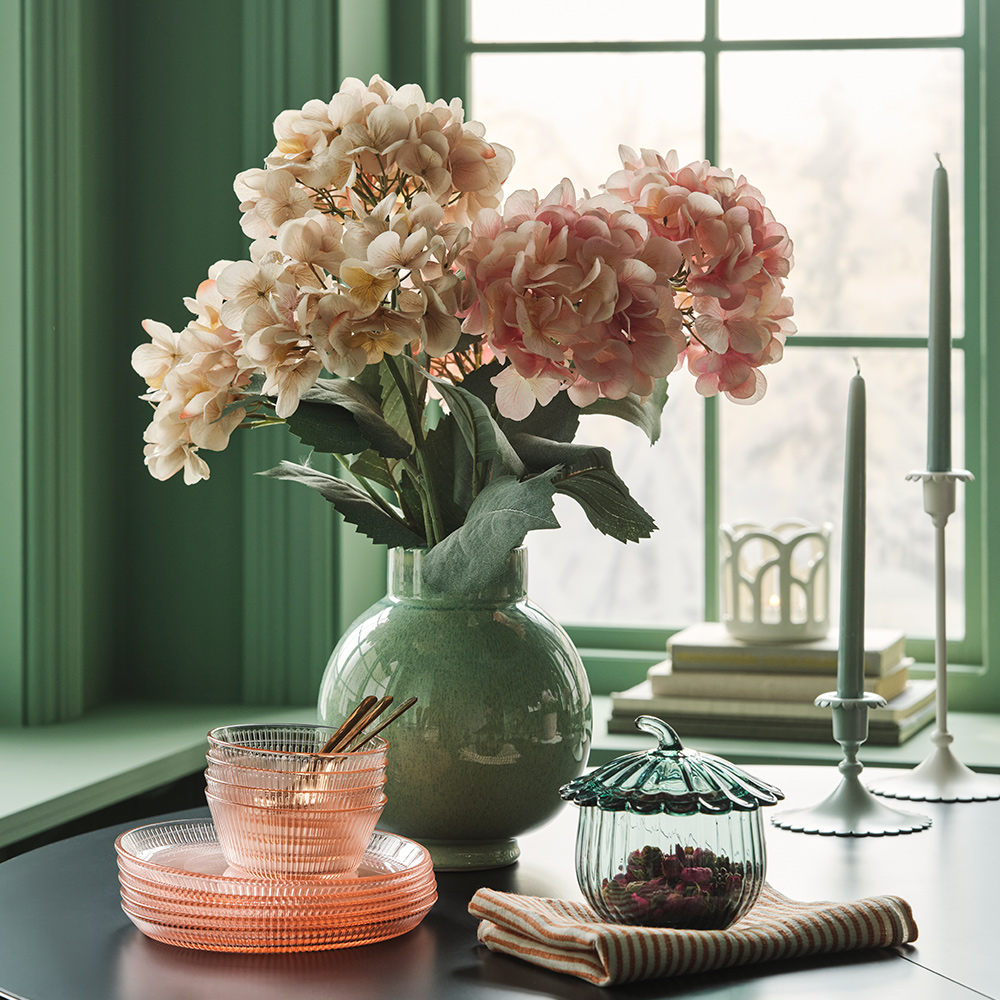 Un ramo de flores artificiales SMYCKA en tonos rosa claro y beige se encuentra en un jarrón verde sobre una mesa con platos y cuencos de cristal.