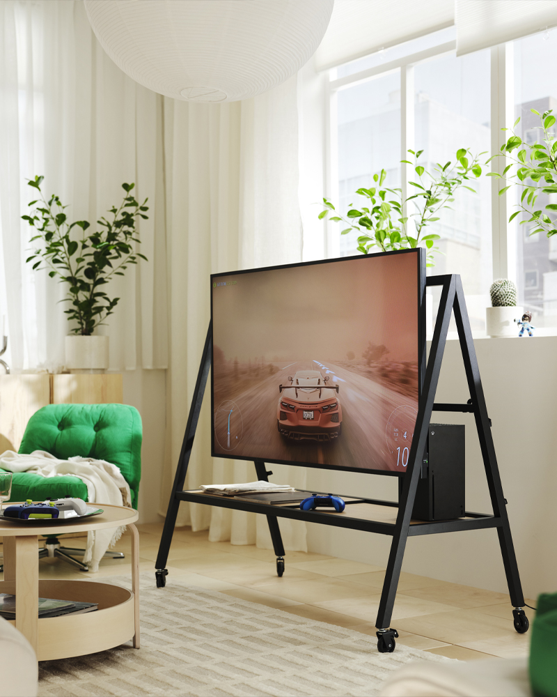 Soporte de TV móvil con ruedas en color negro mostrando un juego de carreras, junto a una consola Xbox en una habitación gamer.