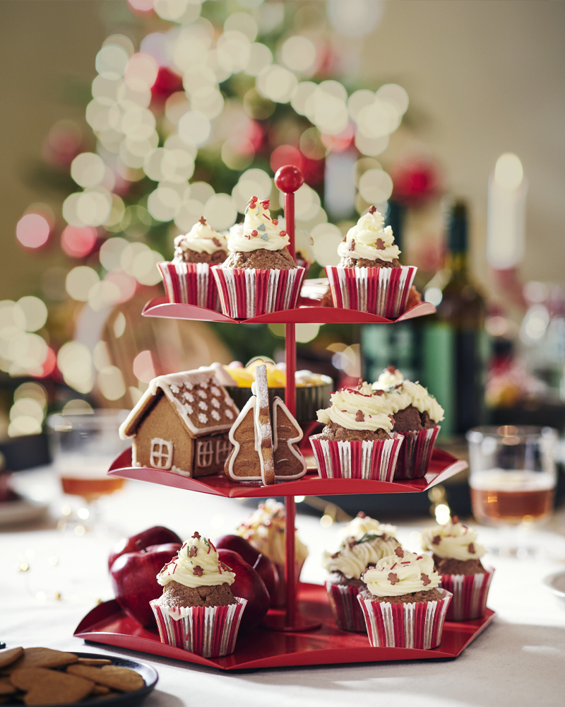 Expositor rojo de 3 niveles con cupcakes navideños y galletas de jengibre, con luces de Navidad desenfocadas de fondo.