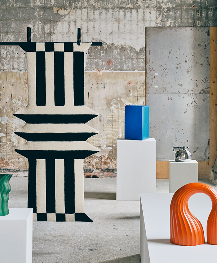 Artículos de la colección KONSTRUNDA de IKEA en una composición de inspiración, con una alfombra a rayas, jarrones y elementos decorativos en negro, azul, naranja y verde sobre pedestales blancos.