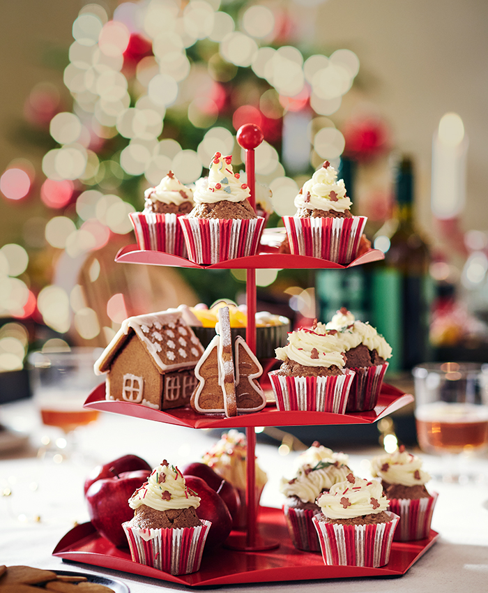 Cupcakes y dulces navideños servidos en una bandeja de varios niveles roja, con un árbol de Navidad decorado al fondo.
