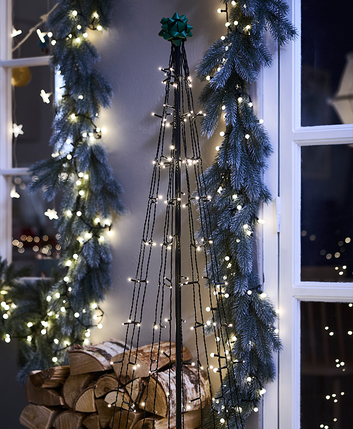 Decoración navideña con luces LED STRÅLA de IKEA, iluminando ramas de abeto y un árbol de Navidad junto a una ventana.