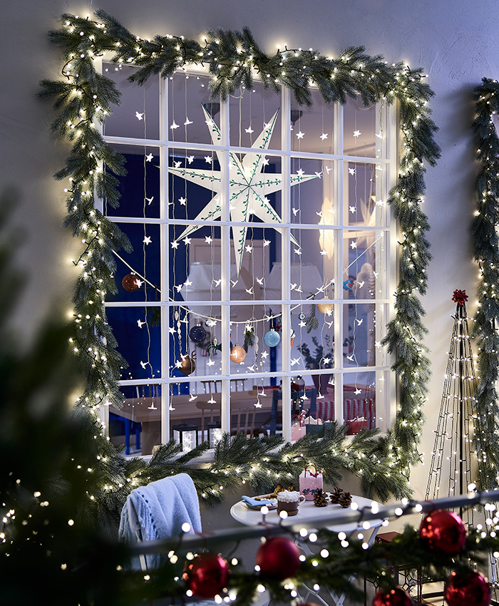 Decoración navideña con luces STRÅLA de IKEA, que iluminan una guirnalda de ramas de abeto alrededor de una ventana decorada con estrellas y adornos navideños.