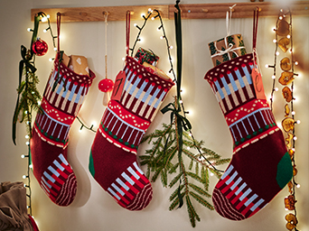 Three red and green Christmas stockings hanging alongside fir branches and decorative lights.