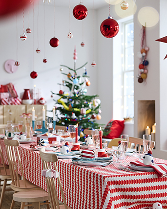 Christmas dining area with a table set with red and white tableware and textiles, hanging Christmas decorations and a Christmas tree in the background.