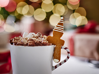 Mug of hot chocolate topped with whipped cream and a Christmas tree-shaped gingerbread biscuit, in a festive setting.