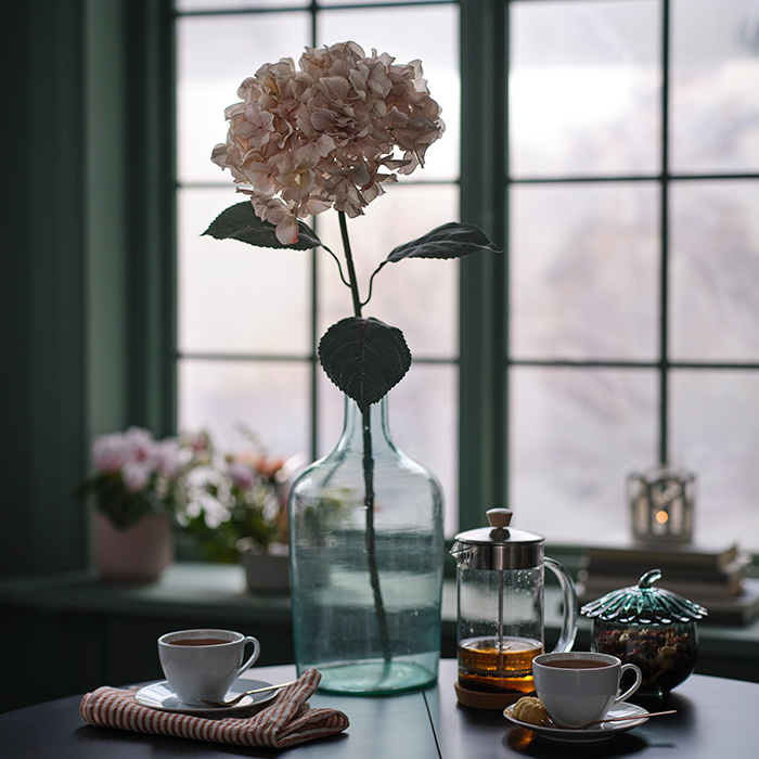 A table with teacups and a bowl of nuts. In a transparent vase in the middle of the table is a SMYCKA artificial flower.