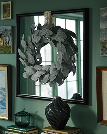 A SMYCKA artificial wreath is hanging on a mirror in a green living room with paintings and books around it.