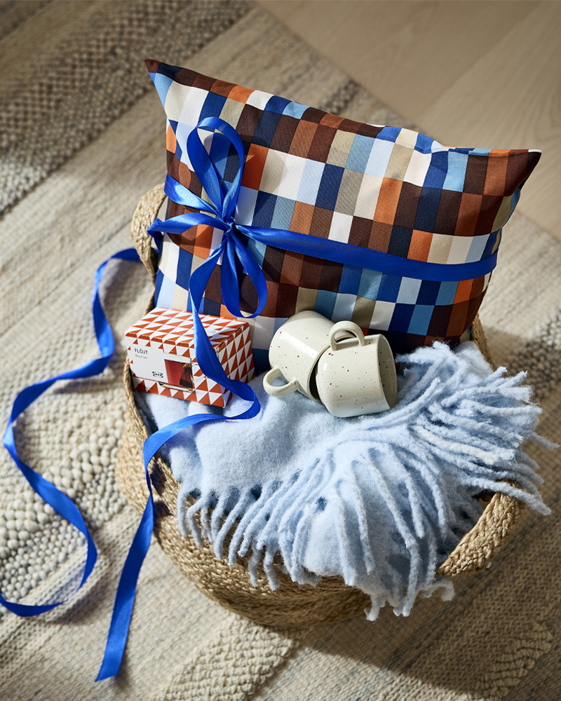 Cesta de mimbre para regalo con cojín a cuadros atado con una cinta azul, manta de manta azul claro y tazas de cerámica.