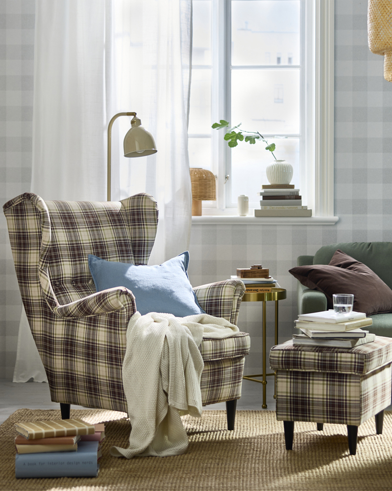 Sillón y reposapiés a cuadros con manta acogedora, lámpara de pie y libros junto a una ventana en el salón.