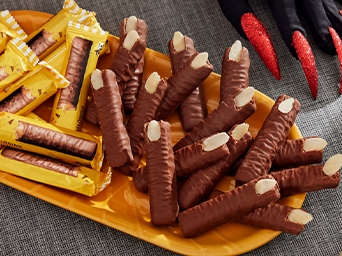 Plato ovalado amarillo con galletas de chocolate en forma de dedos con almendra por uña, junto a barritas empaquetadas y manos de bruja.