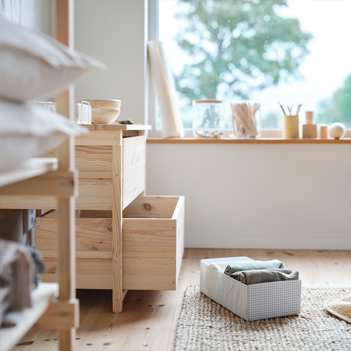 Bright, sunlit room featuring a solid pine wooden drawer unit next to a grey grid-pattern fabric organizer with folded clothes resting on a jute rug.