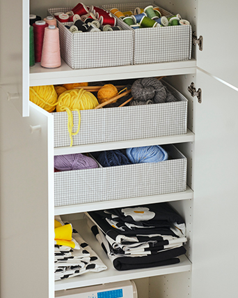 White wardrobe interior organized with light grey checked fabric boxes containing colorful sewing threads, yarn skeins, and neatly folded patterned textiles.