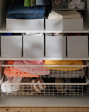 Closet interior with white shelves and wire pull-out baskets, holding grey grid-pattern fabric organizers with neatly stacked jeans, folded clothes, and rolled garments.