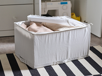 Light grey grid-pattern fabric storage box with a zippered mesh lid partially open, storing folded blankets and garments on a black and white striped rug.