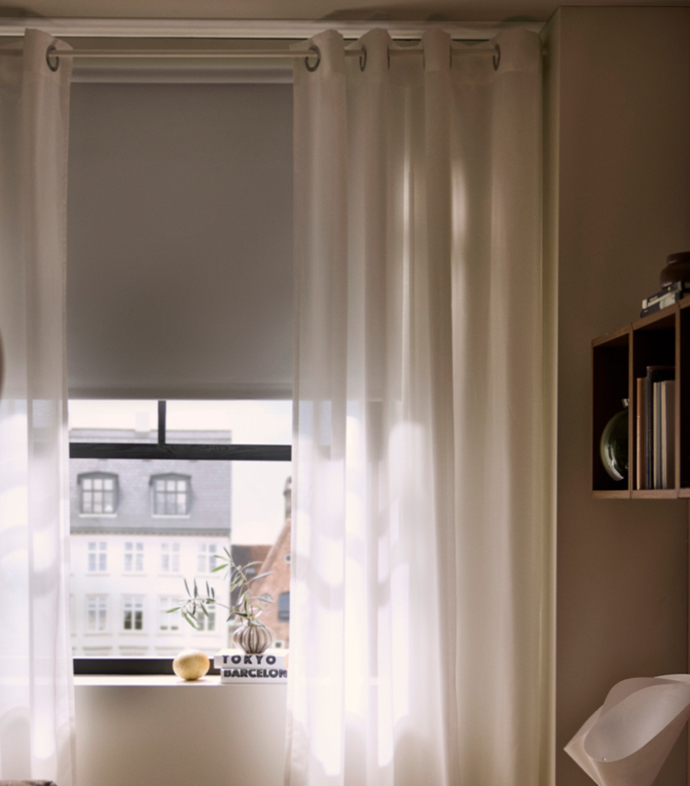 Window with a grey roller blind framed by sheer white eyelet curtains, featuring a shelf unit on the right and city view outside.